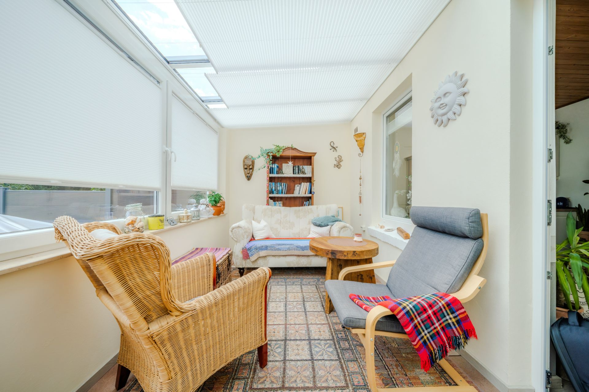 Heller Wintergarten mit zwei Sesseln, Sofa, Holztisch und Regal mit Büchern.