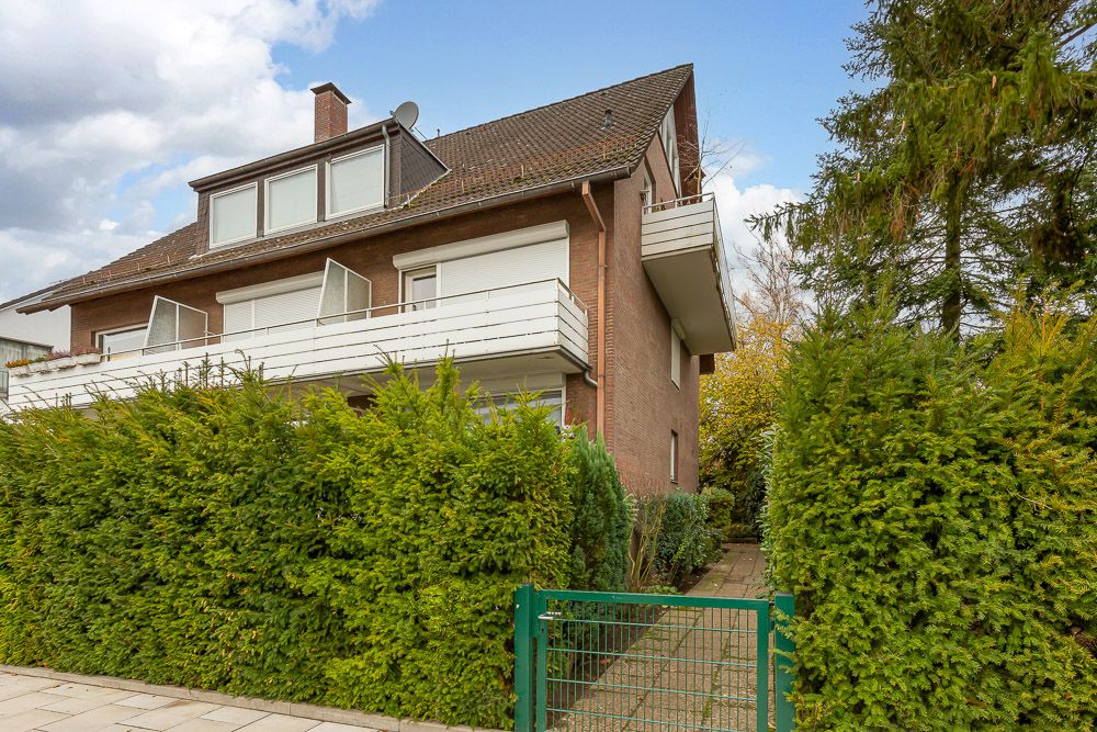 Mehrfamilienhaus mit Klinkerfassade, Balkon und grünem Vorgarten mit Zaun.