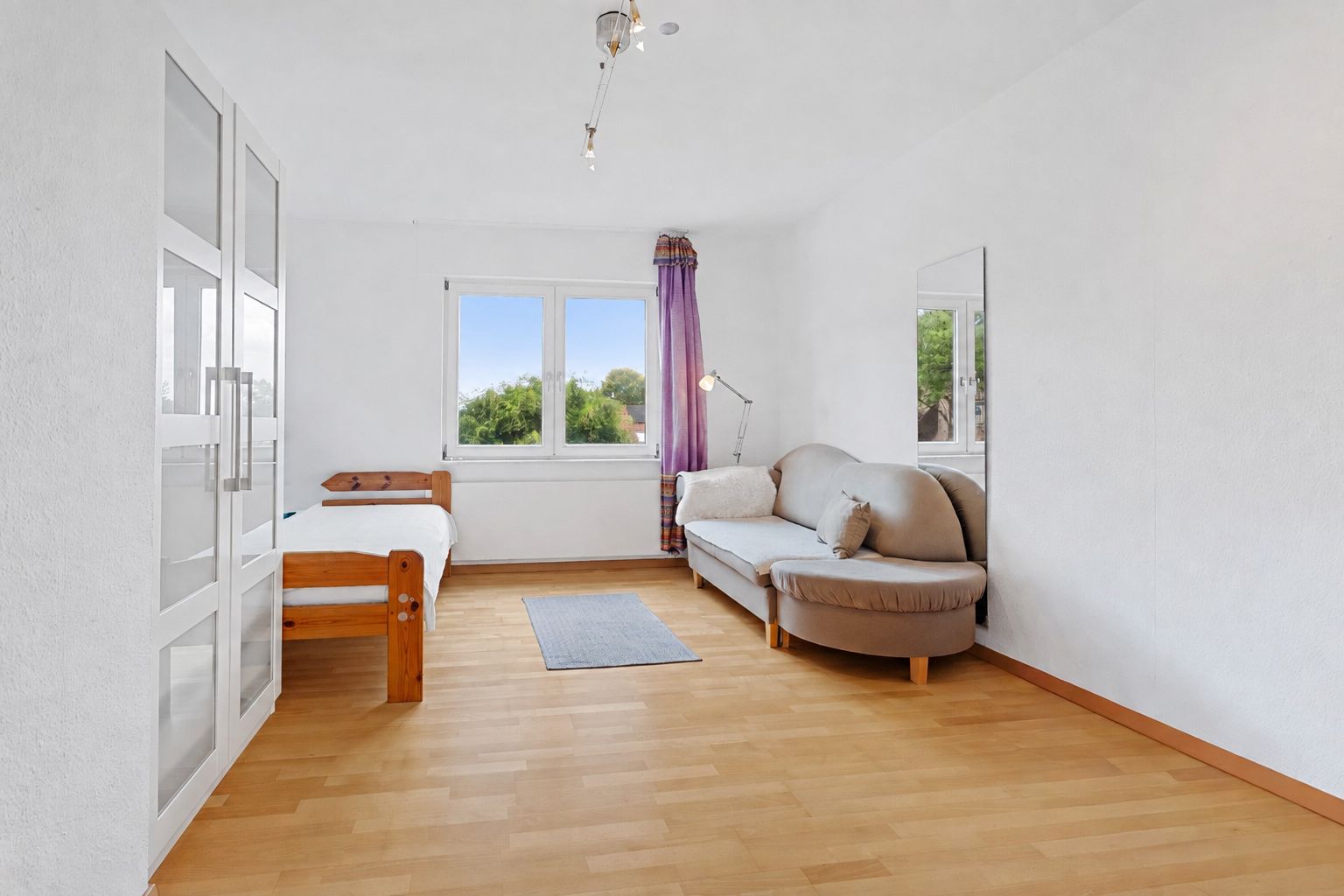 Helles Zimmer mit Holzbett, Sofa, Teppich, Kleiderschrank und Fenster mit lila Vorhang.