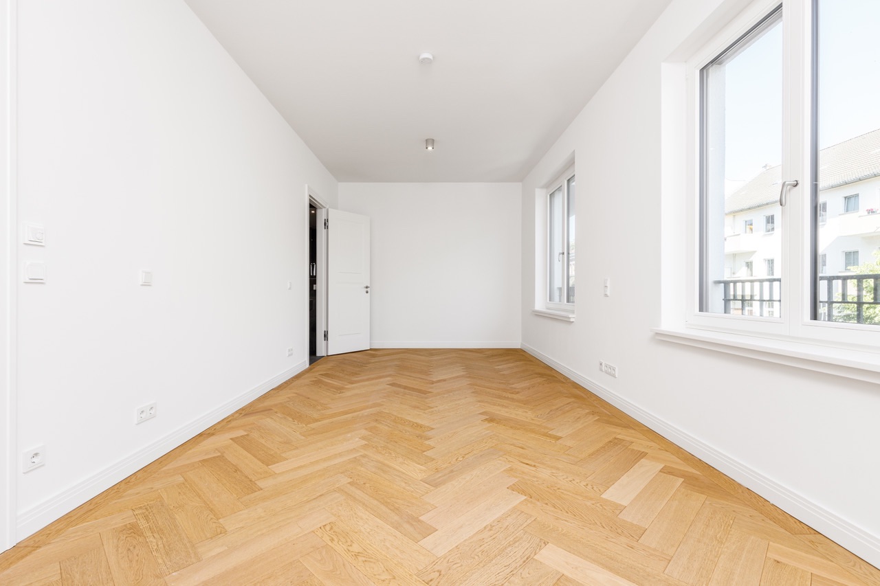 Leeres Zimmer mit Fischgrätparkett, weißen Wänden und zwei großen Fenstern.