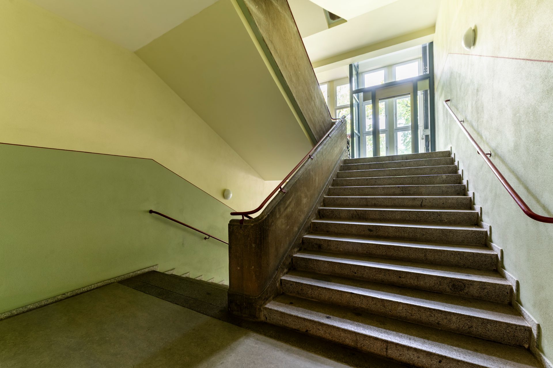 Treppenhaus mit breiter Steintreppe und Handläufen, Tageslicht durch große Glastür am oberen Ende.