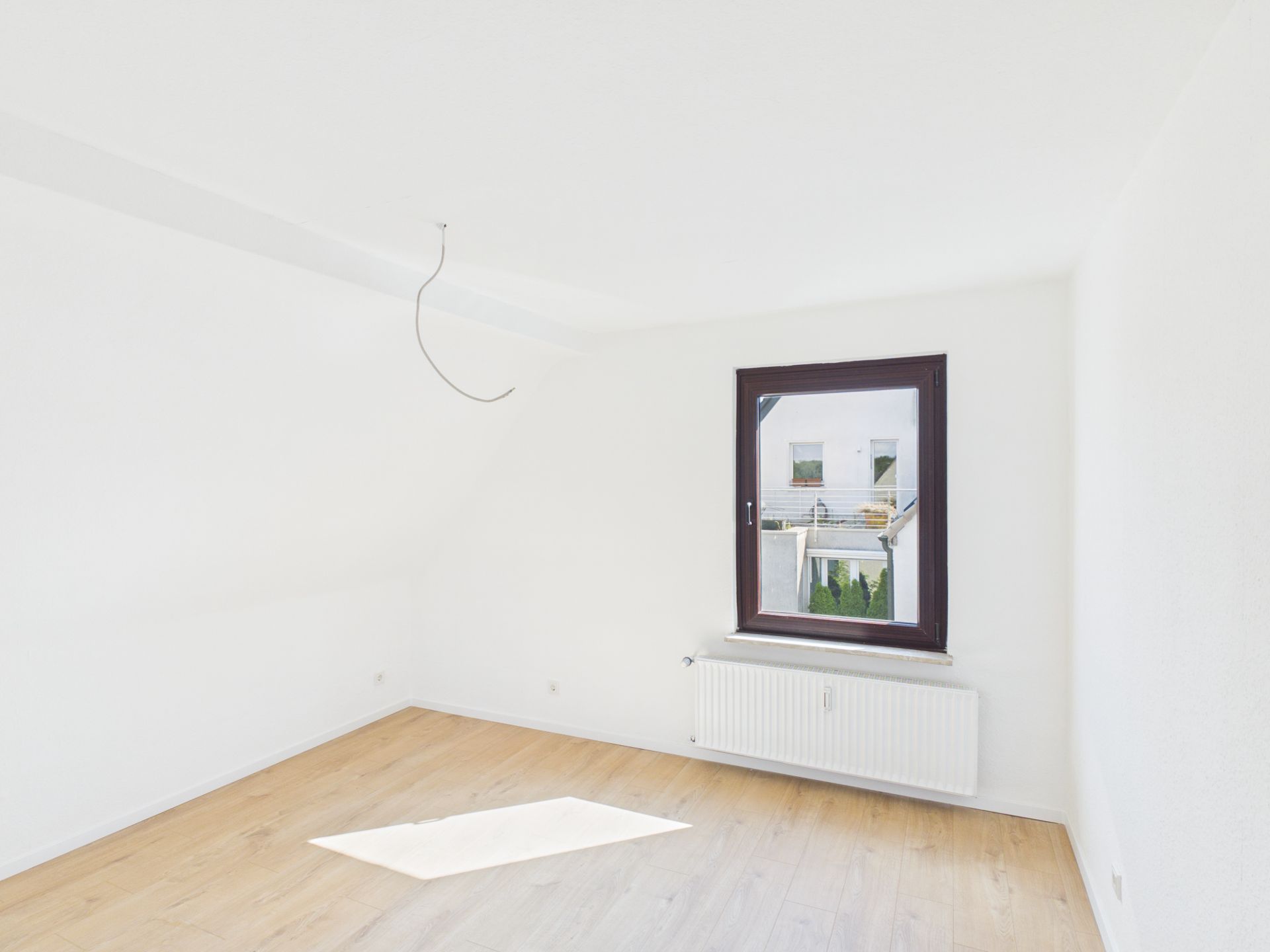 Helles, leeres Zimmer mit Holzfußboden, weißer Wand und einem Fenster mit Heizkörper darunter.
