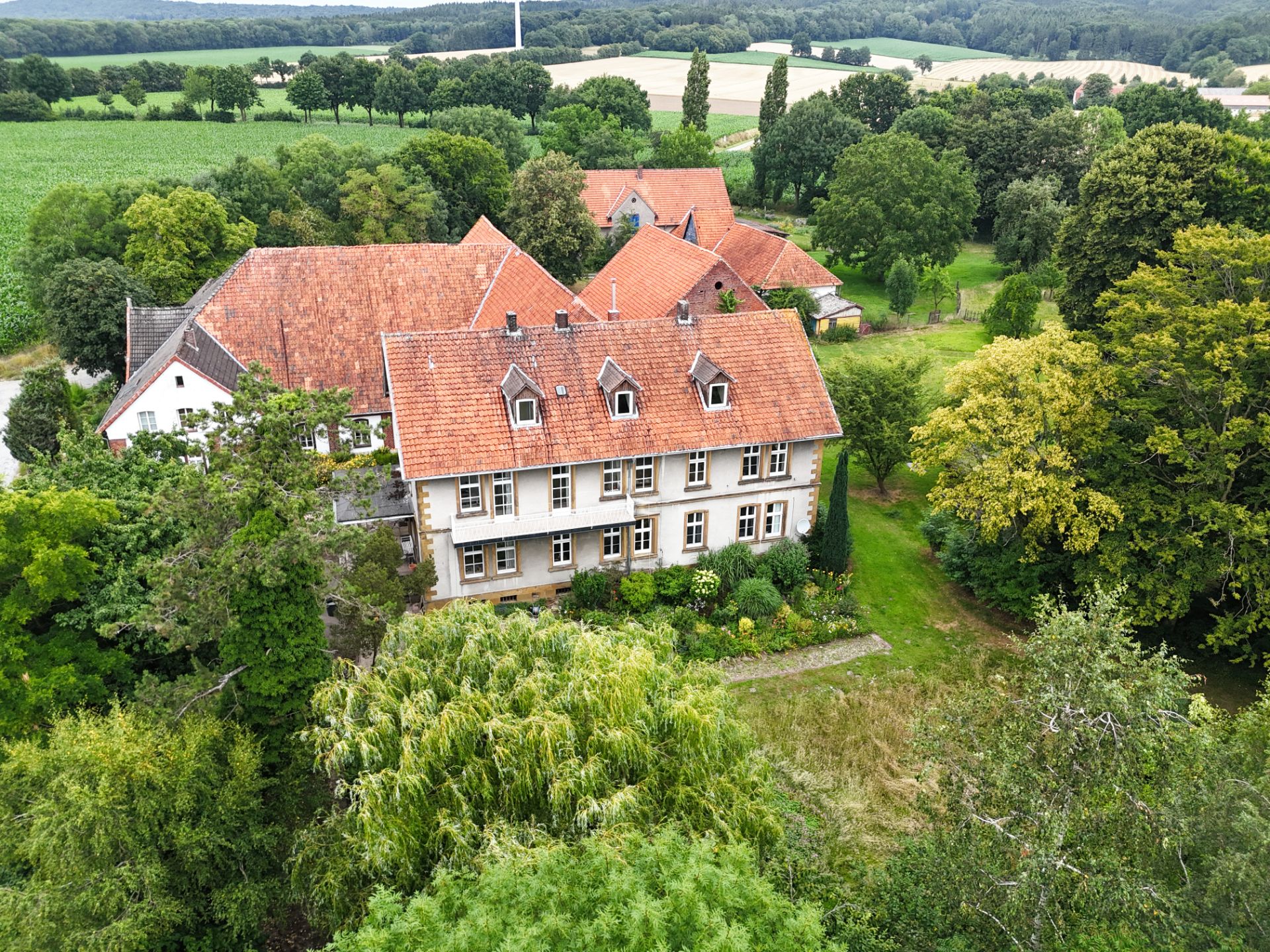 Großes Wohnhaus mit rotem Dach, umgeben von Bäumen und Garten, im ländlichen Gebiet.