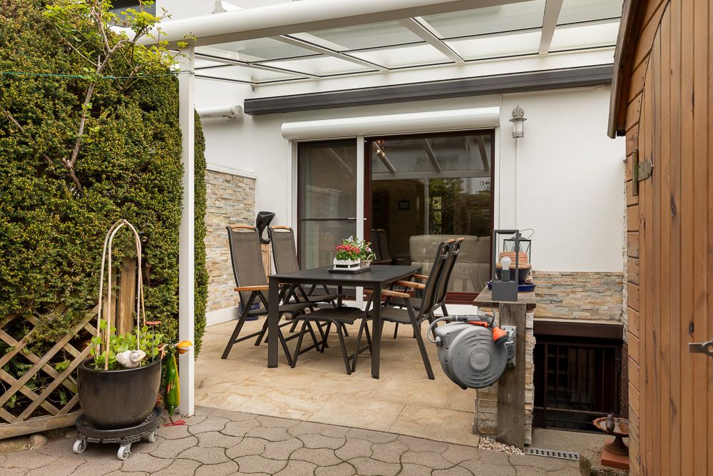 Überdachte Terrasse mit Esstisch, vier Stühlen und Pflanzen neben einem Wohnhaus.