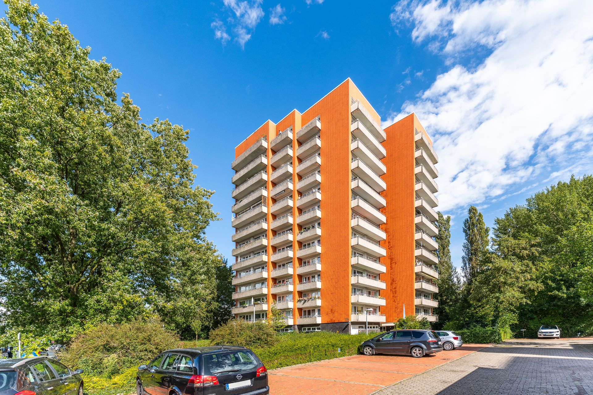 Hohes Wohngebäude mit orangefarbener Fassade, Balkonen und Parkplatz, umgeben von Bäumen.