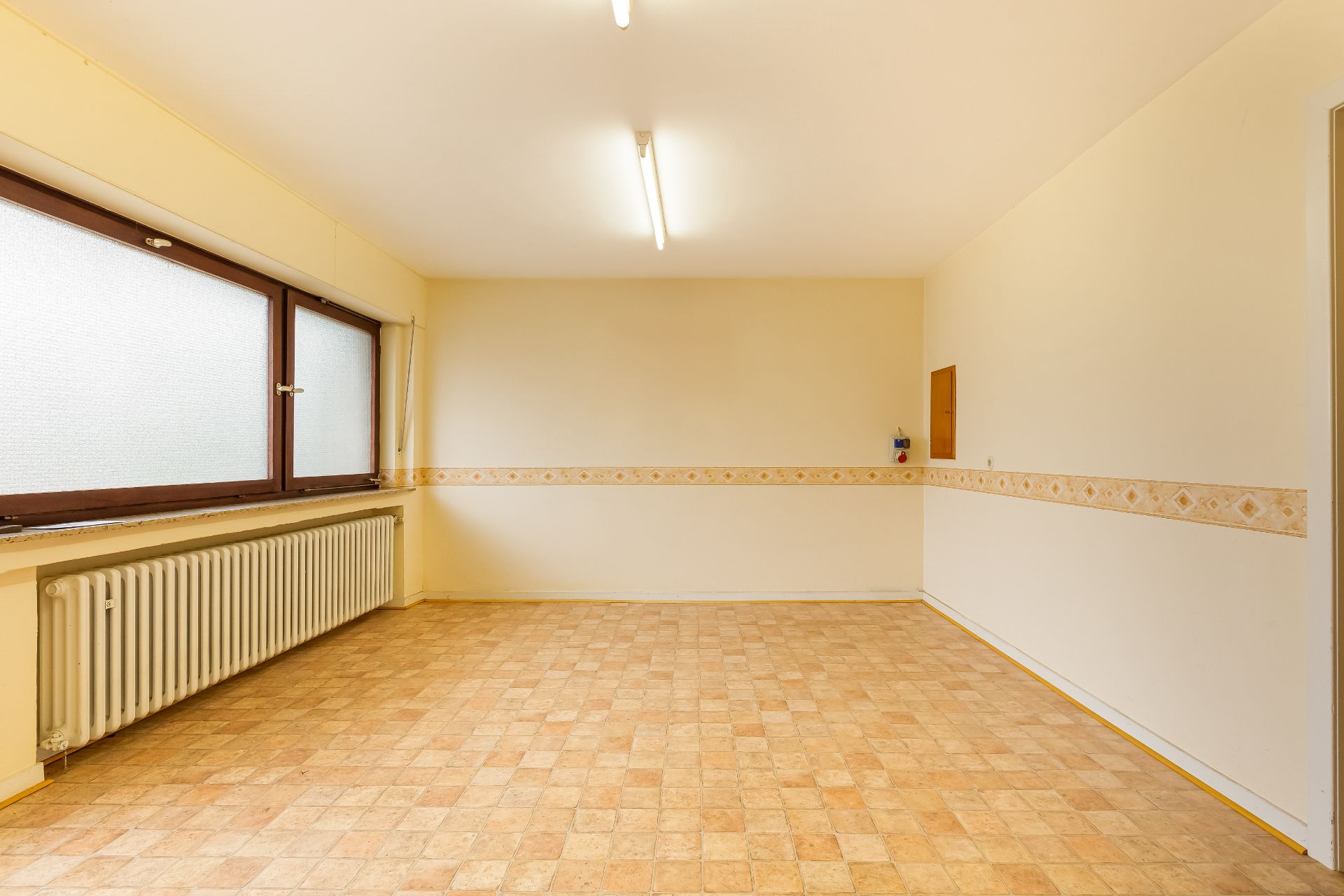 Leerer Raum mit großen Fenstern, Heizung und gemustertem Streifen an der Wand.