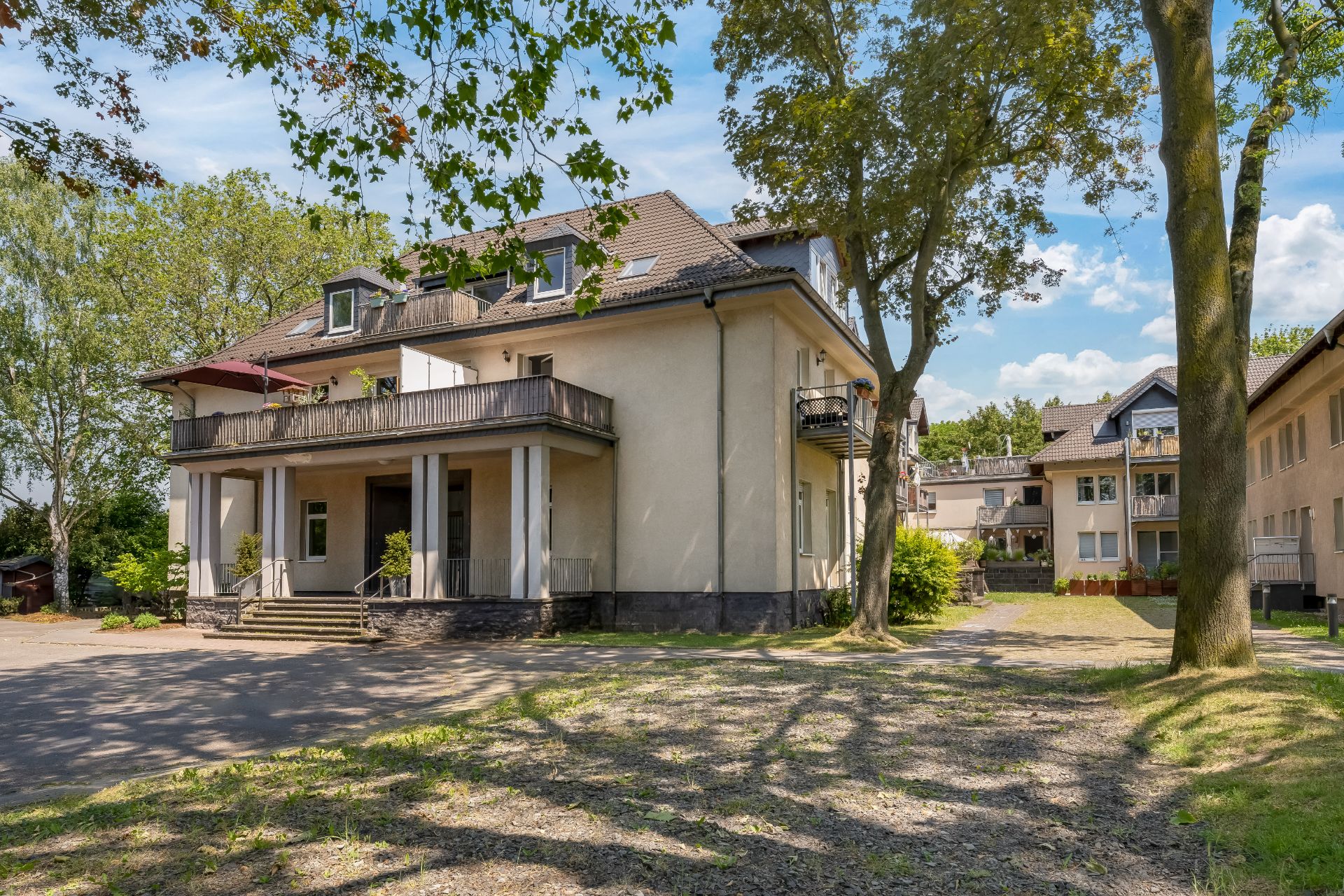 Zweistöckiges Wohnhaus mit großem Balkon, Treppe zum Eingang und Bäumen im Vordergrund.