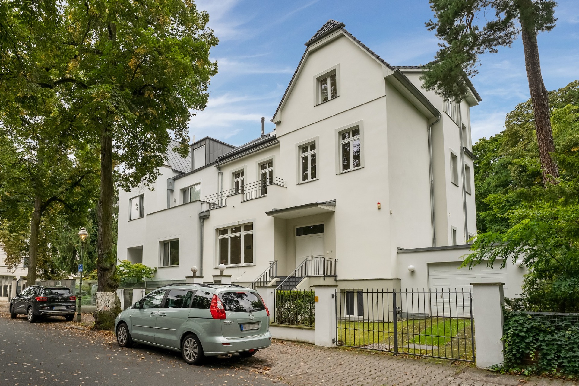 Großes weißes Wohnhaus mit mehreren Fenstern, Vorgarten und parkenden Autos an der Straße.