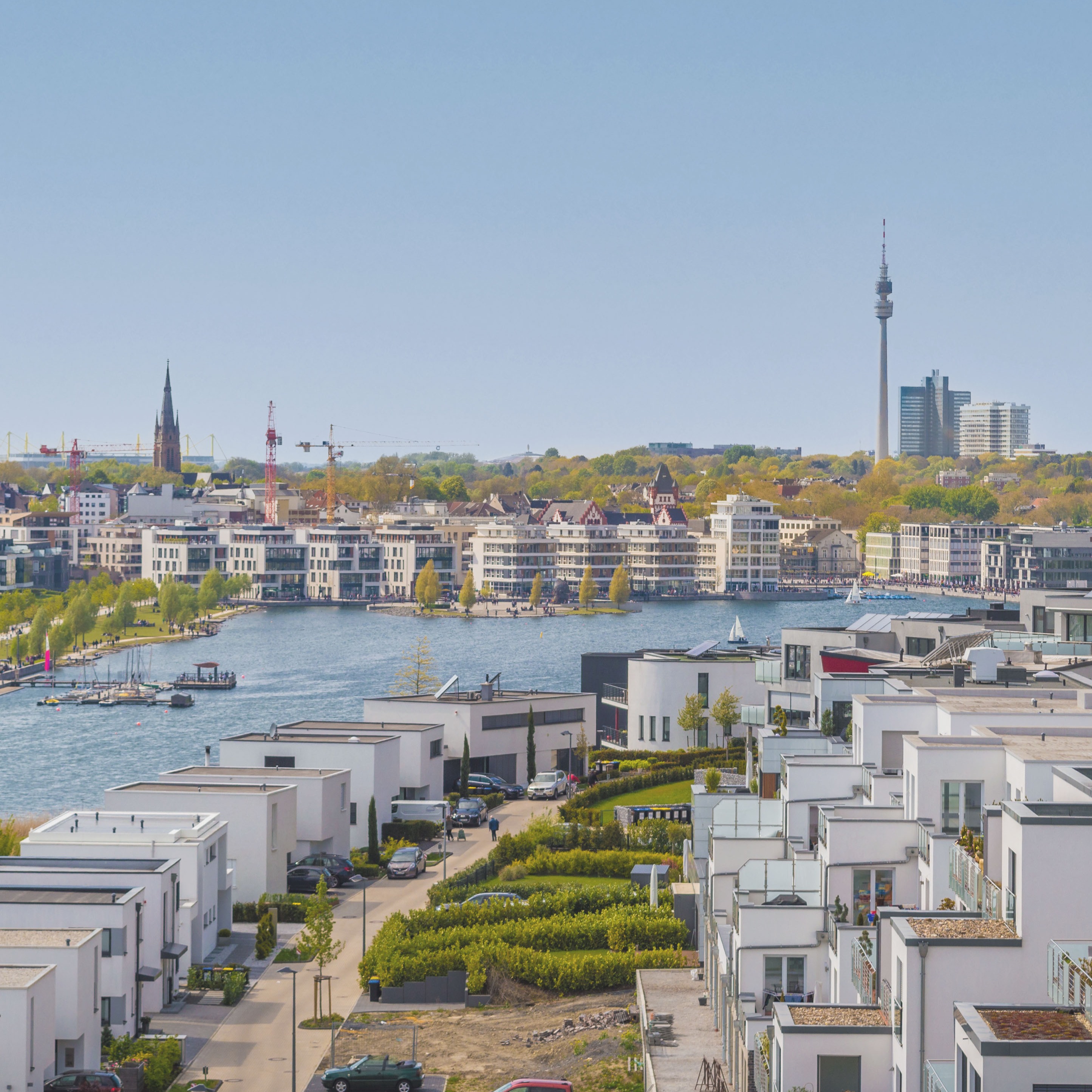 Moderne Wohnhäuser am Wasser mit Blick auf Stadt, Fernsehturm und Segelboote bei klarem Himmel.
