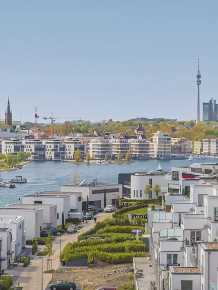 Moderne Wohnhäuser am Wasser mit Blick auf Stadt, Fernsehturm und Segelboote bei klarem Himmel.