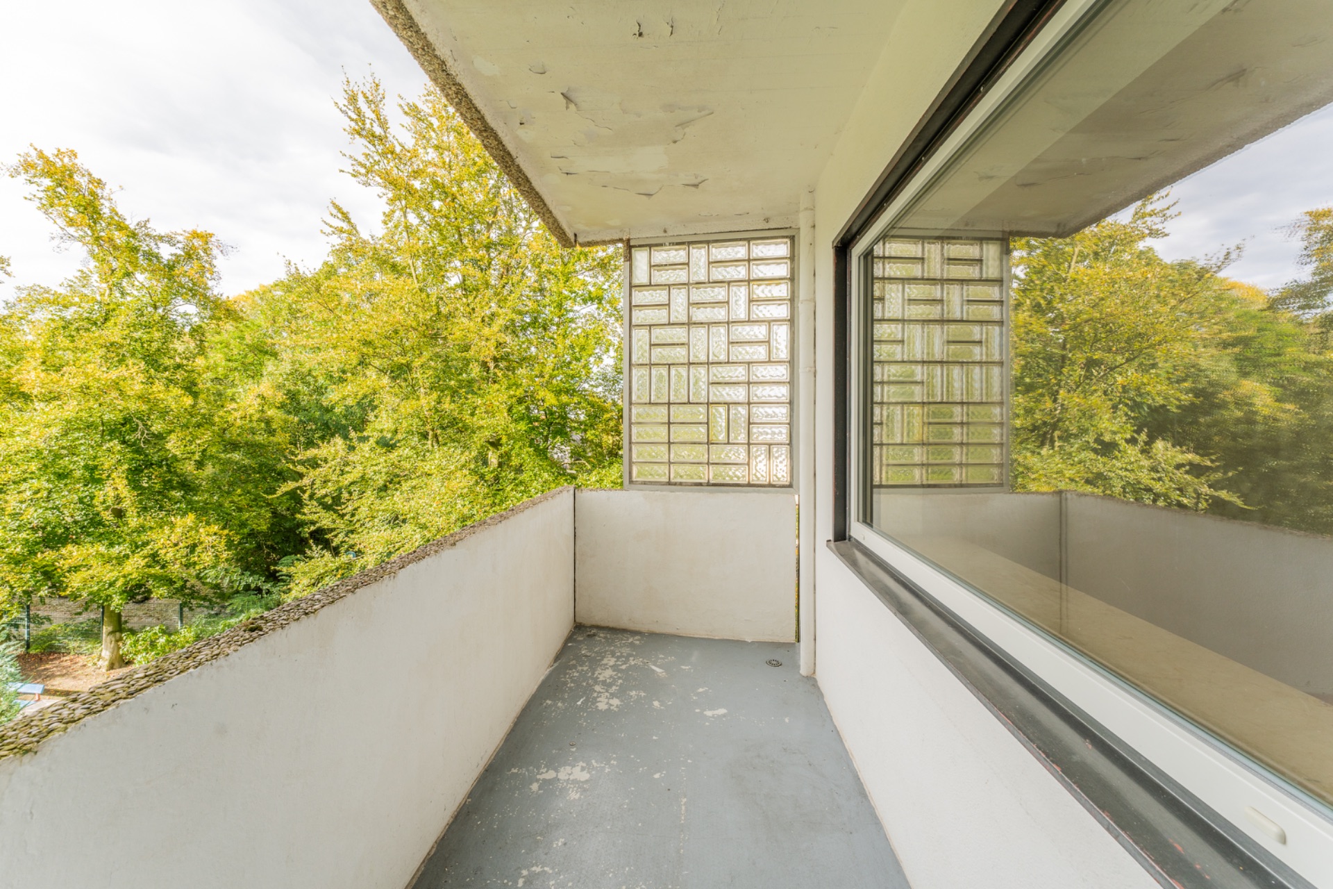 Balkon mit weißer Brüstung und Glasbausteinwand, Blick auf grüne Bäume.