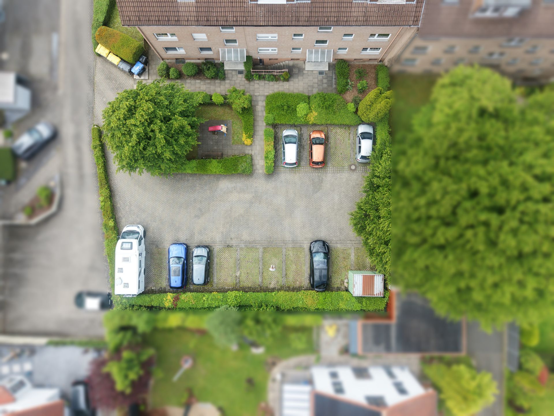 Luftaufnahme eines Parkplatzes vor einem Wohngebäude mit mehreren geparkten Autos und viel Grün.
