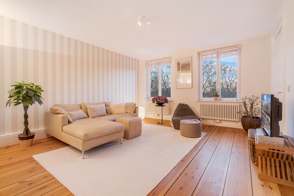Helles Wohnzimmer mit beiger Eckcouch, Sessel, Pflanzen und Parkettboden.