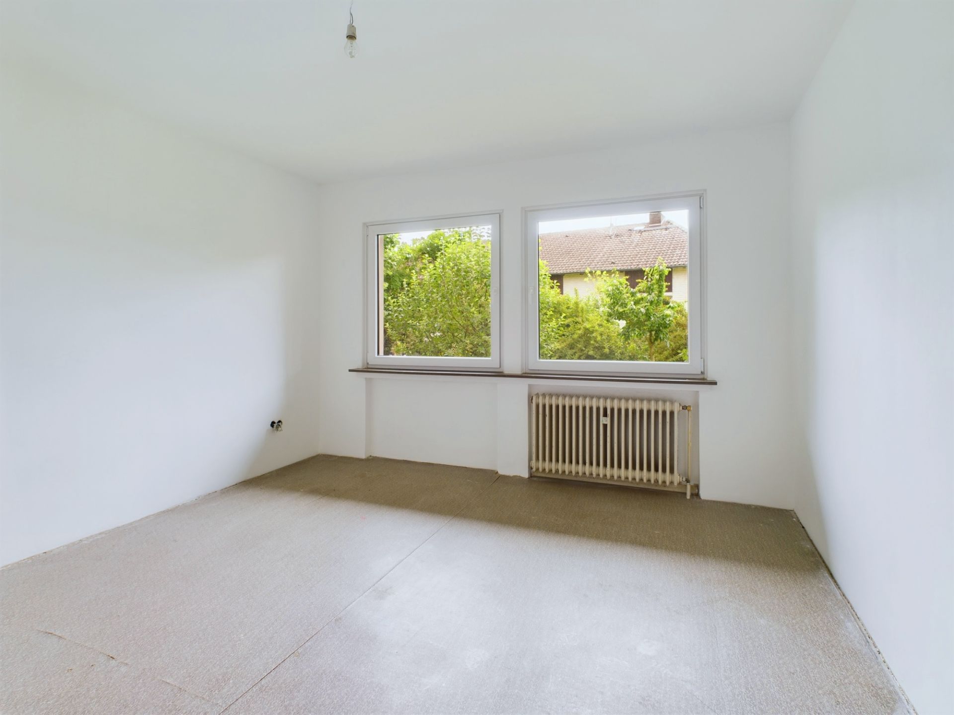 Leerer Raum mit weißen Wänden, zwei großen Fenstern und einem Heizkörper unter den Fenstern.