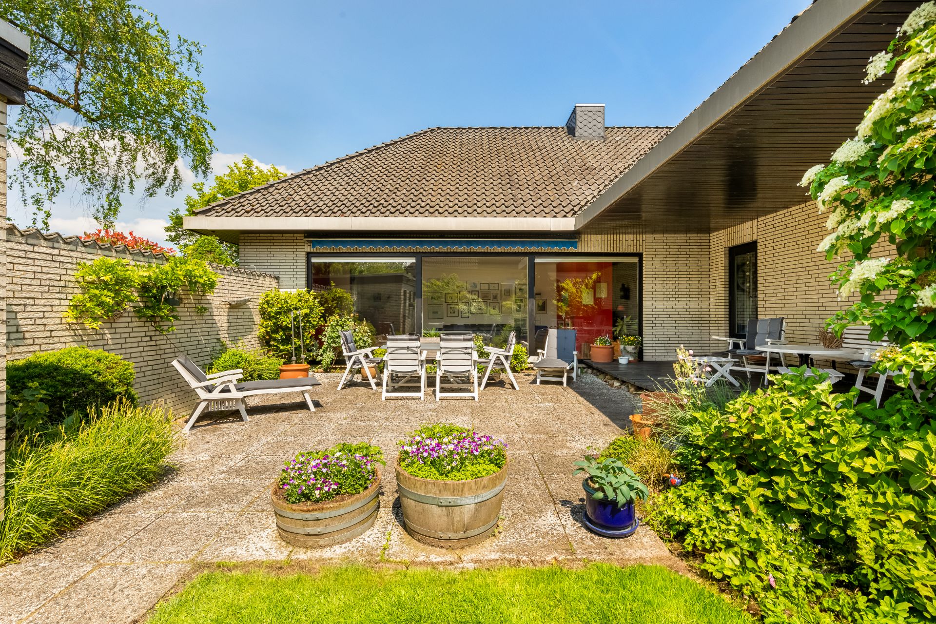 Terrasse mit Gartenmöbeln, Blumen und Pflanzen vor einem Einfamilienhaus bei sonnigem Wetter.