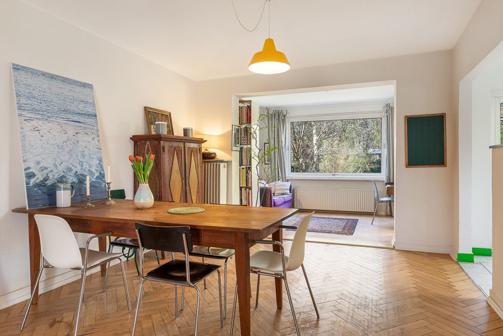 Esszimmer mit Holztisch, vier Stühlen, Blumen, großem Fenster und Blick ins Wohnzimmer.