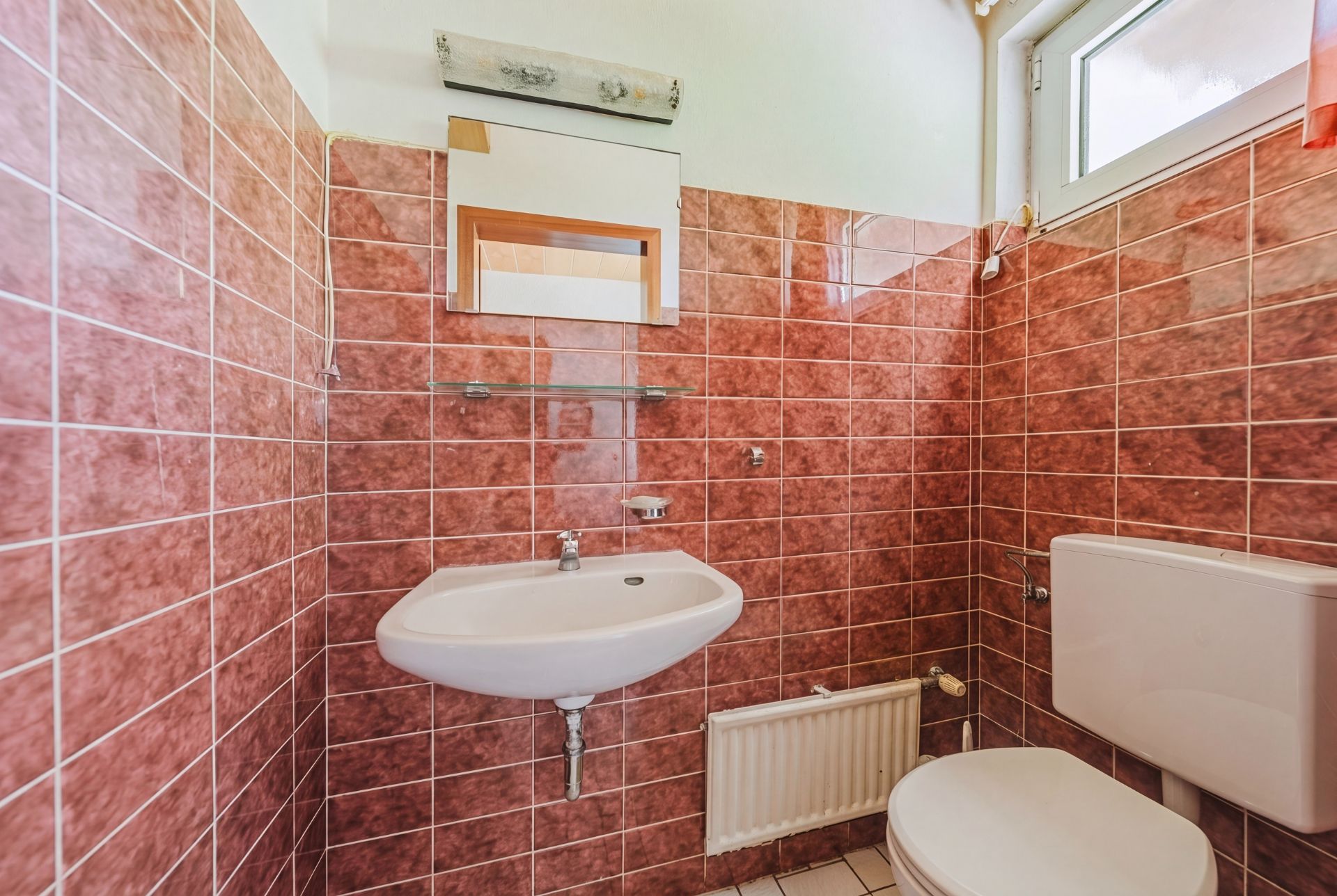 Kleines Bad mit braunen Fliesen, Waschbecken, Spiegel und Toilette.