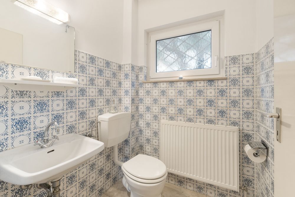 Kleines Bad mit Waschbecken, Toilette, Spiegel und blau-weißen Fliesen, Fenster und Heizkörper.