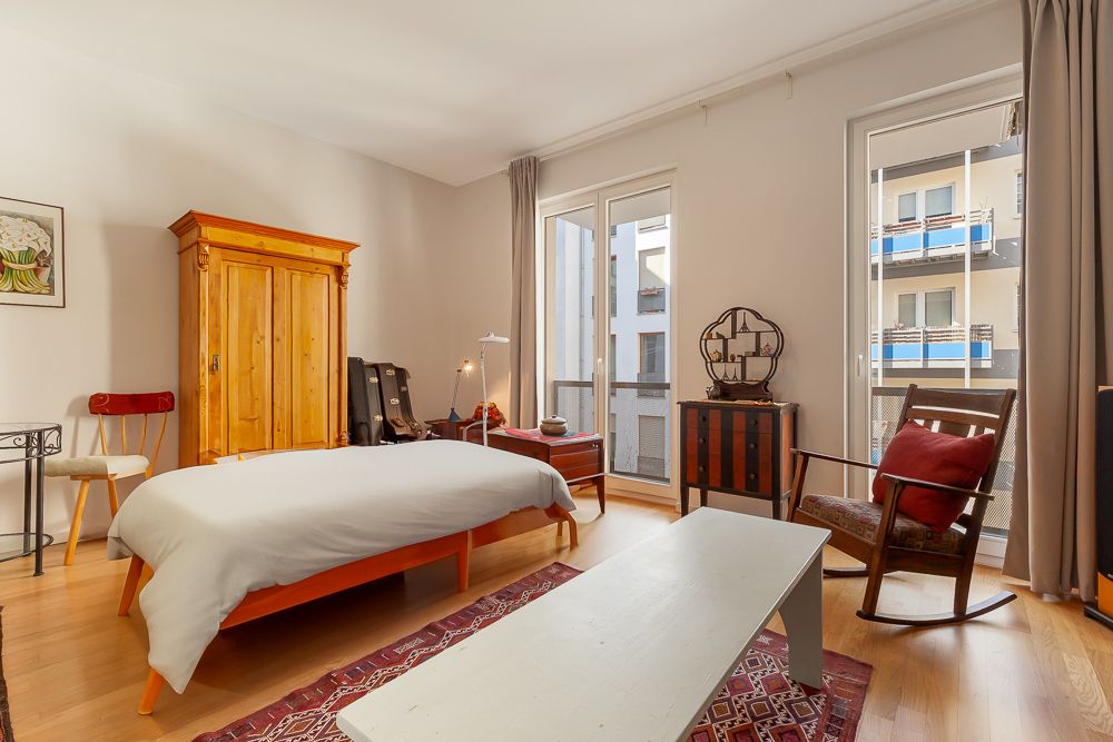 Helles Schlafzimmer mit Holzbett, Schrank, Schaukelstuhl und großem Fenster zu einem Balkon.