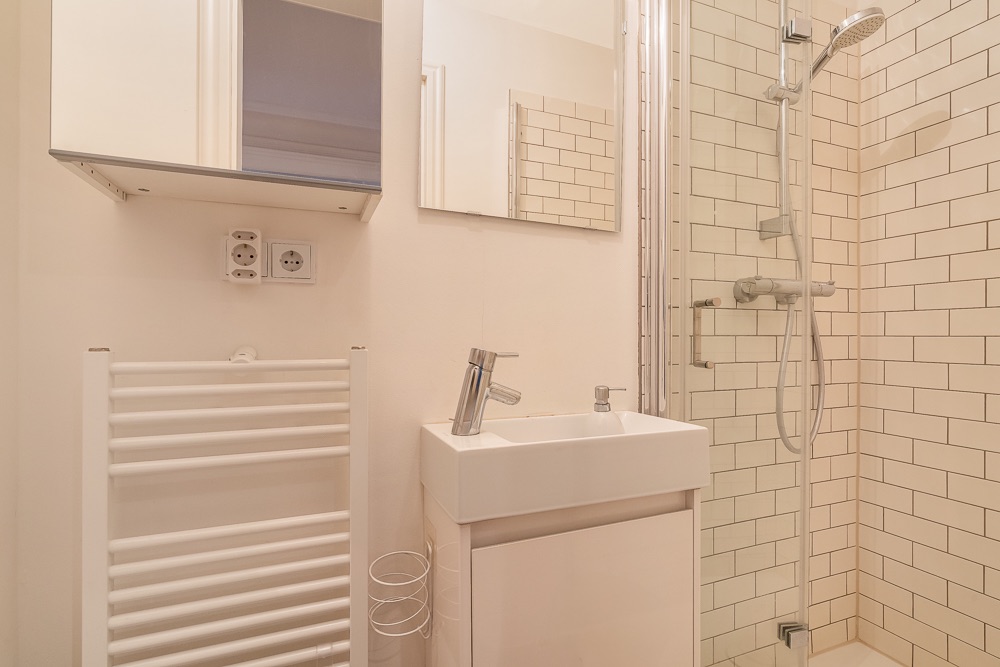 Helles Bad mit Waschbecken, Spiegel, Handtuchheizkörper und Dusche mit weiß gefliester Wand.