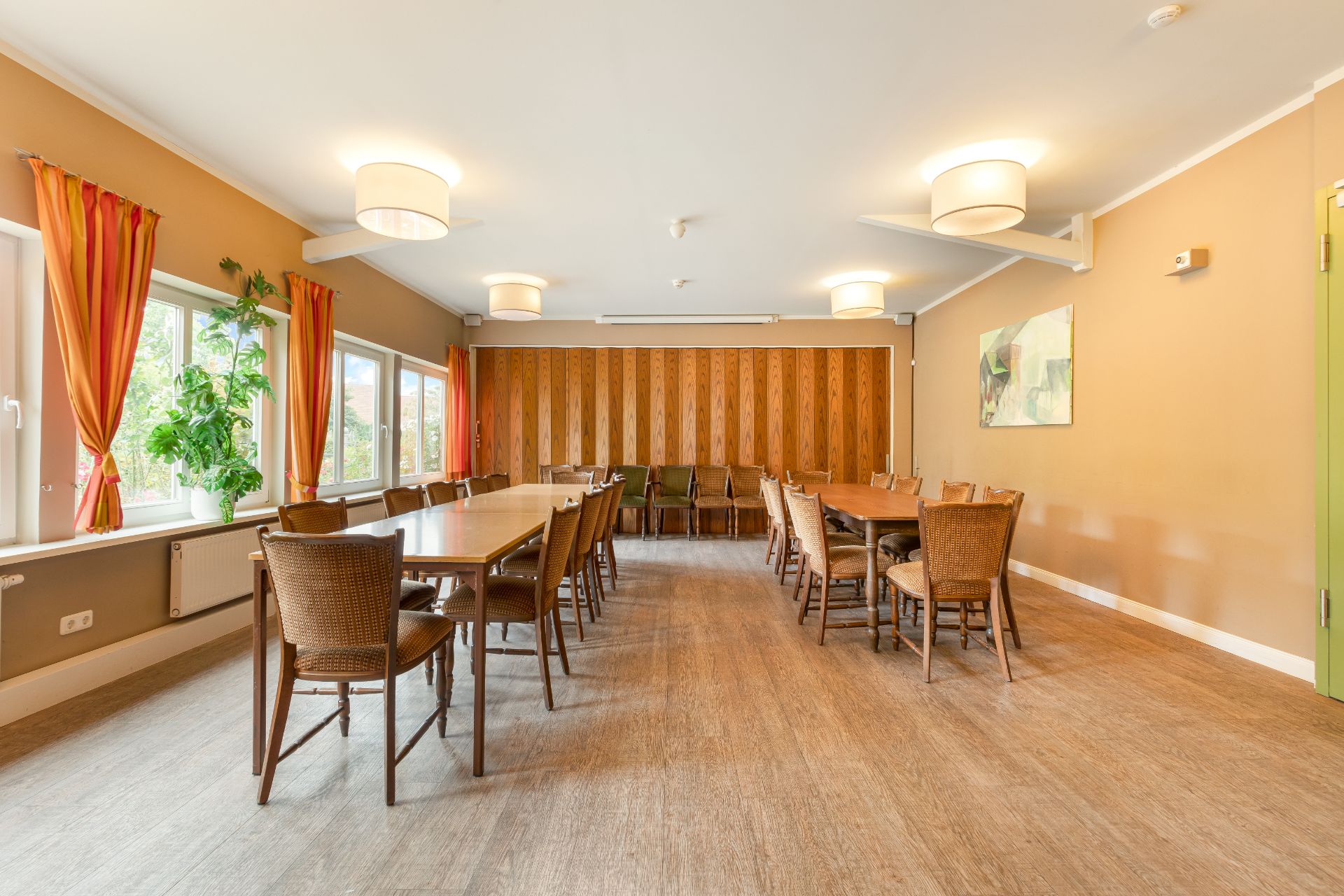 Heller Raum mit langen Holztischen, vielen Stühlen, großen Fenstern und orangefarbenen Vorhängen.