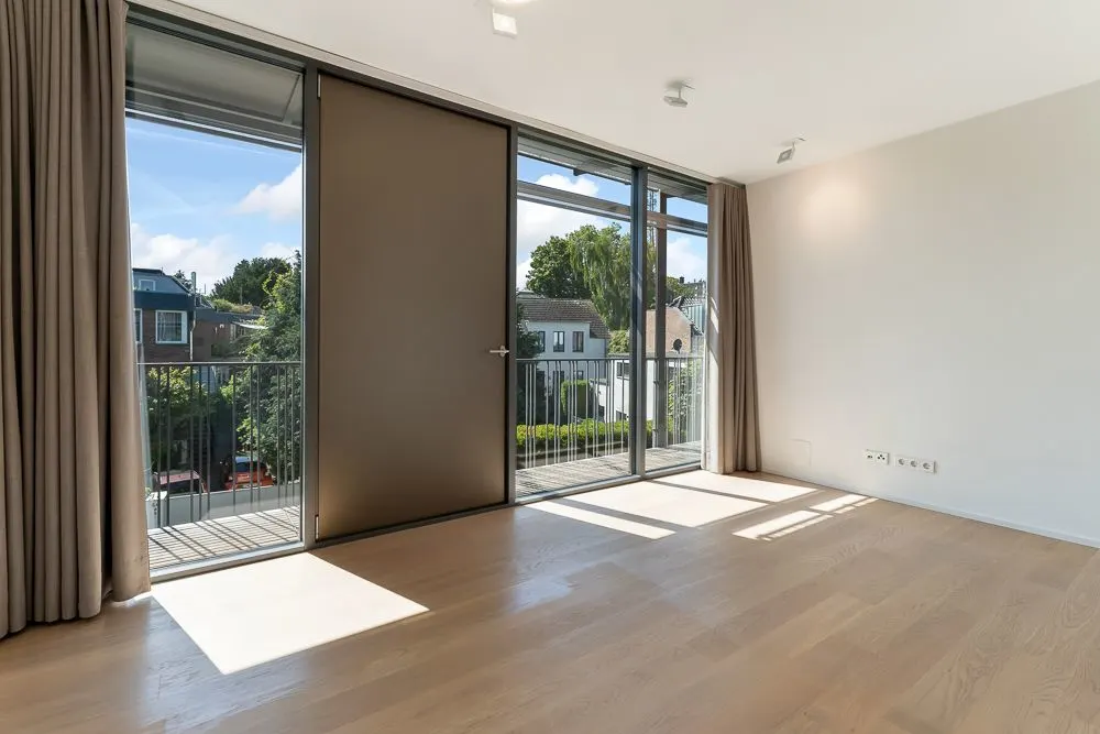 Helles, leeres Zimmer mit großen Fenstern, Holzboden und Zugang zu einem Balkon.