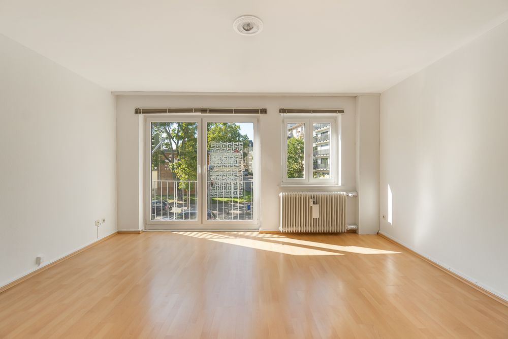 Heller, leerer Raum mit Holzfußboden, großen Fenstern und einem Heizkörper unter dem rechten Fenster.
