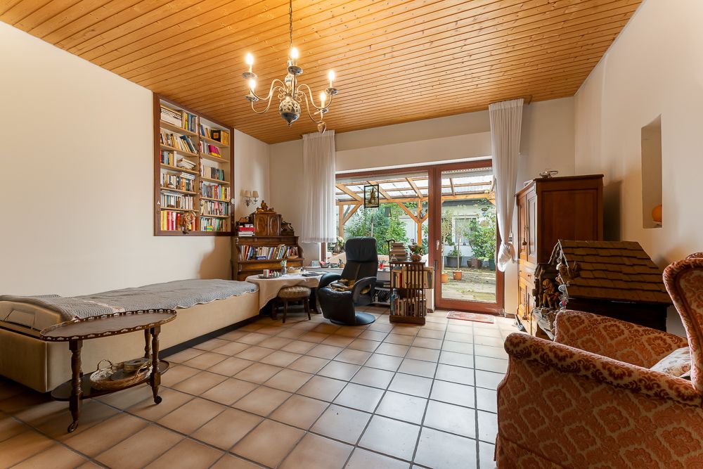 Wohnzimmer mit Fliesenboden, Holzmöbeln, Bücherregal und Blick auf Terrasse mit Garten.