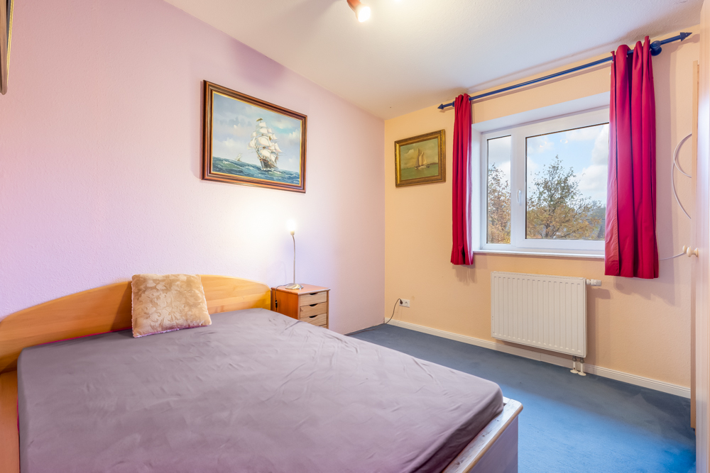 Schlafzimmer mit Doppelbett, Nachttisch, Gemälden an der Wand und Fenster mit roten Vorhängen.