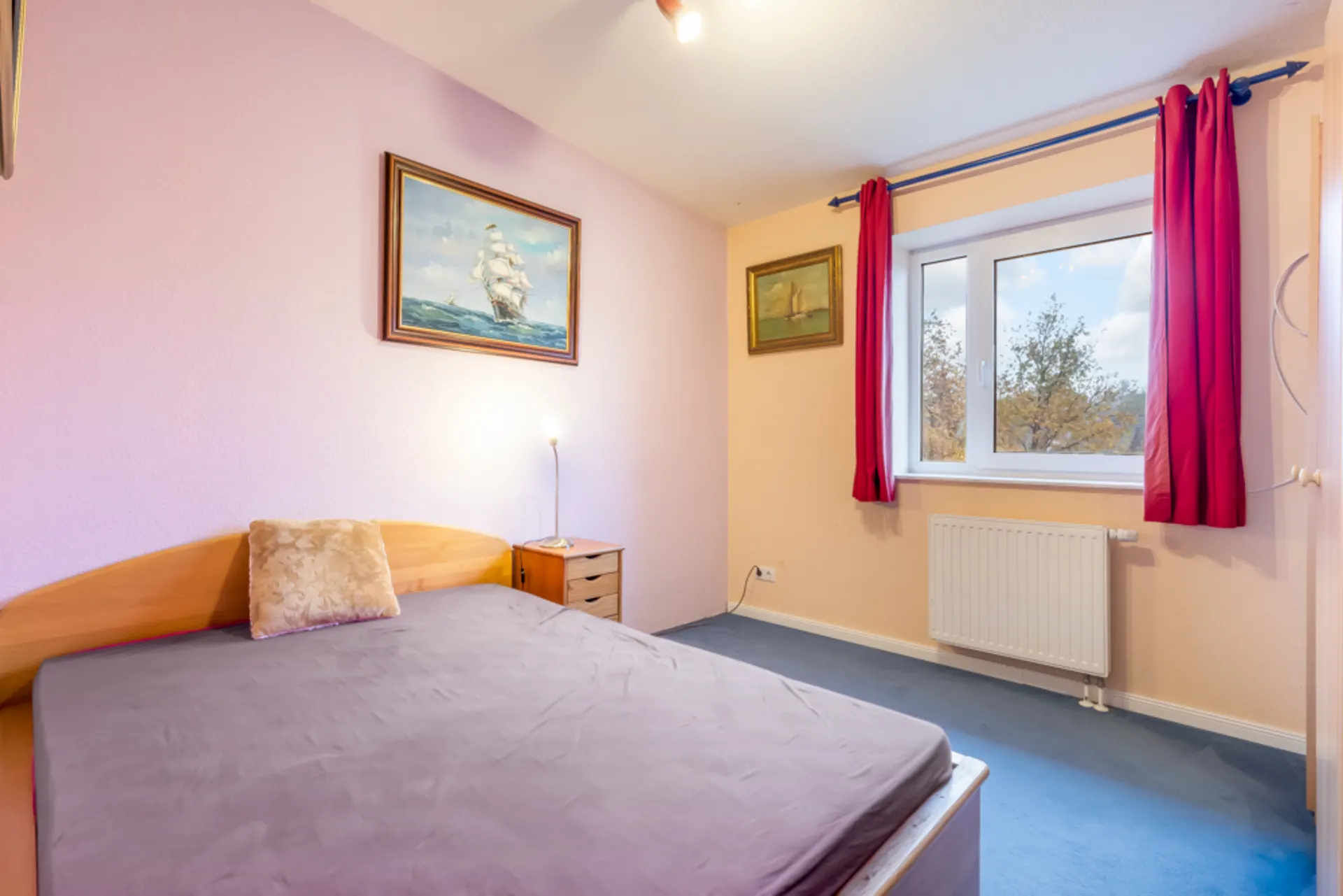 Schlafzimmer mit Doppelbett, Nachttisch, Gemälden an der Wand und Fenster mit roten Vorhängen.