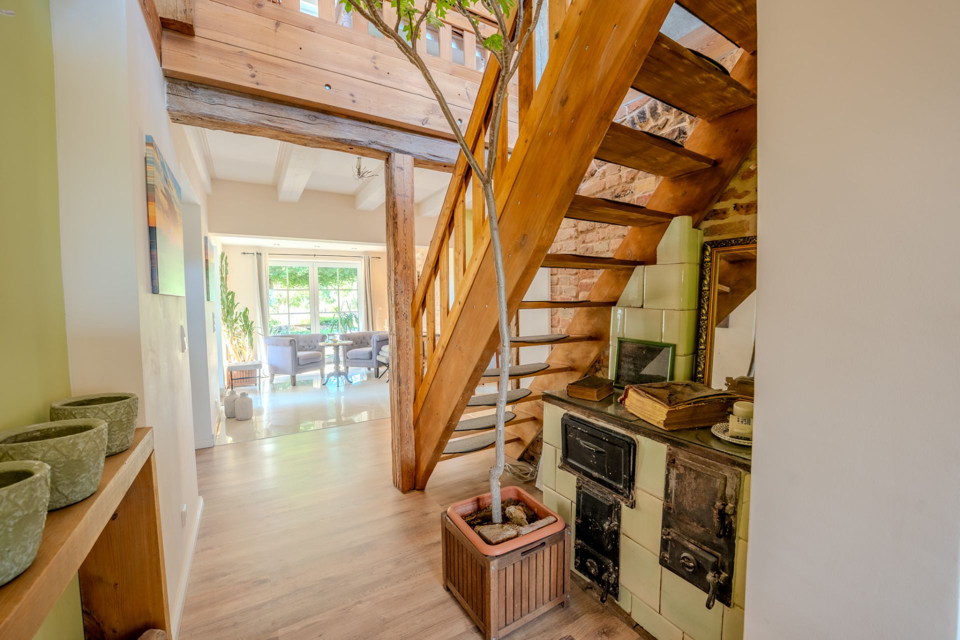 Heller Flur mit Holztreppe, altem Ofen, Topfpflanze und Blick ins Wohnzimmer mit großen Fenstern.