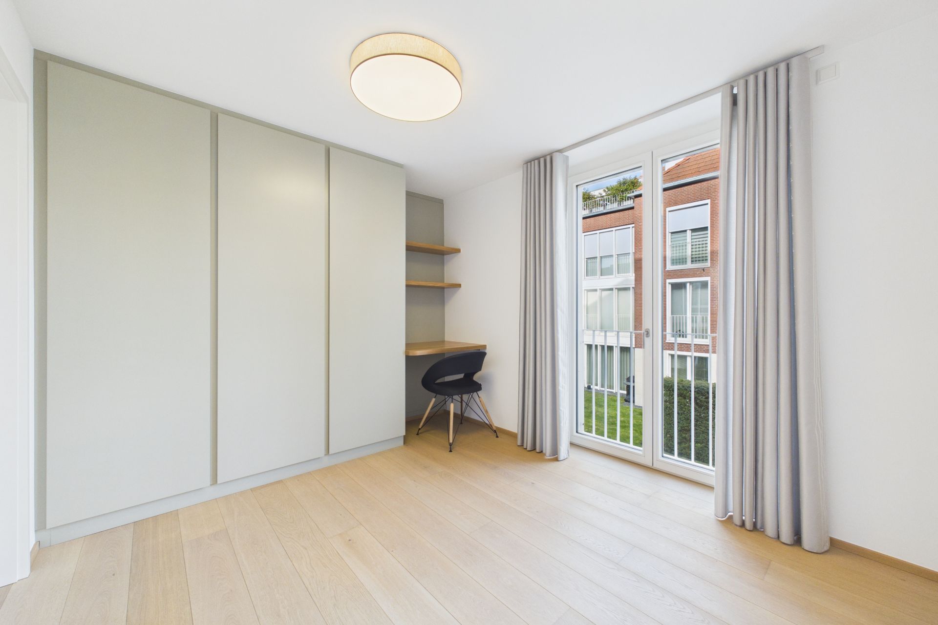 Helles Zimmer mit Einbauschrank, Schreibtisch, Stuhl und großem Fenster mit Vorhängen.