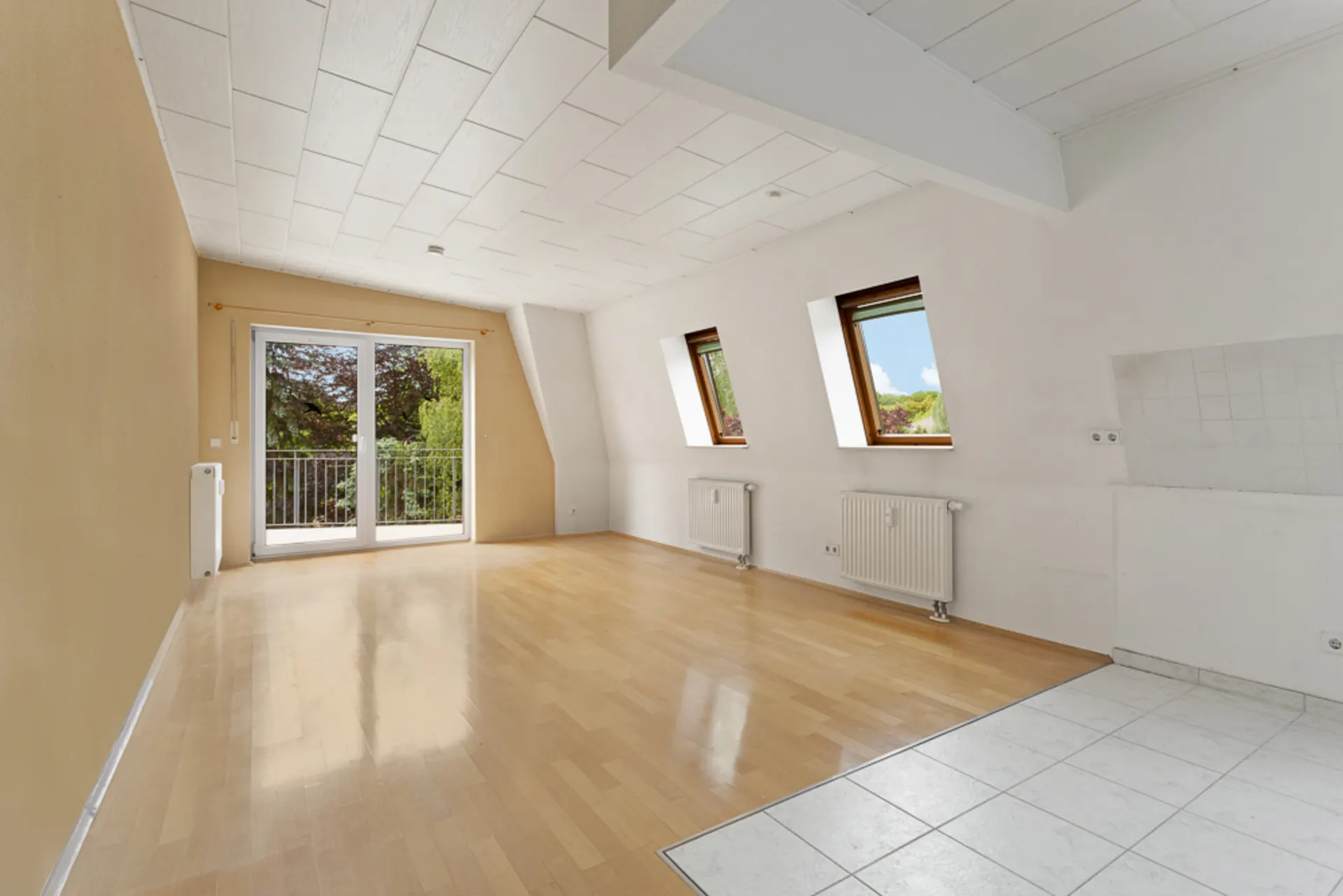 Leerer Raum mit Parkettboden, zwei Fenstern, Balkon und Fliesenbereich neben weißer Wand.