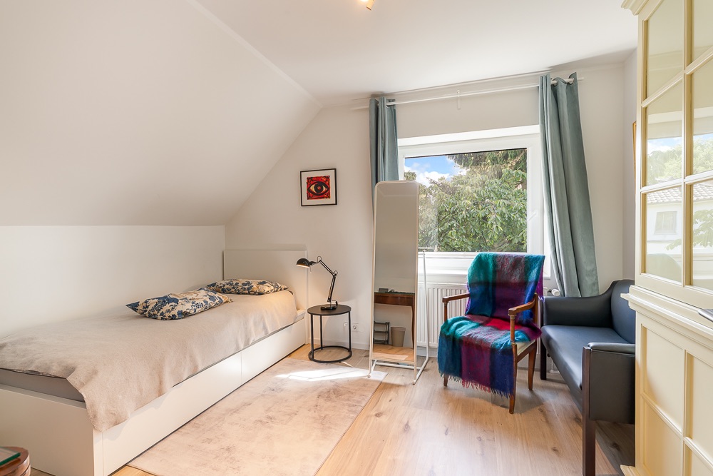 Helles Schlafzimmer mit Einzelbett, Sessel, Stehlampe, Spiegel und großem Fenster mit Vorhängen.