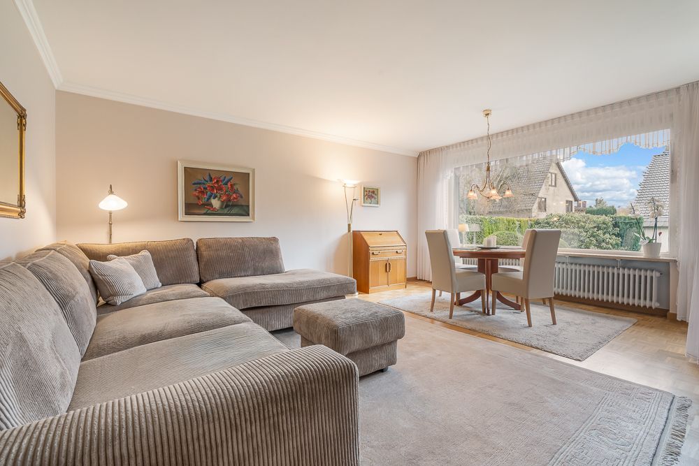 Wohnzimmer mit grauem Ecksofa, Esstisch, großen Fenstern und Bildern an der Wand.