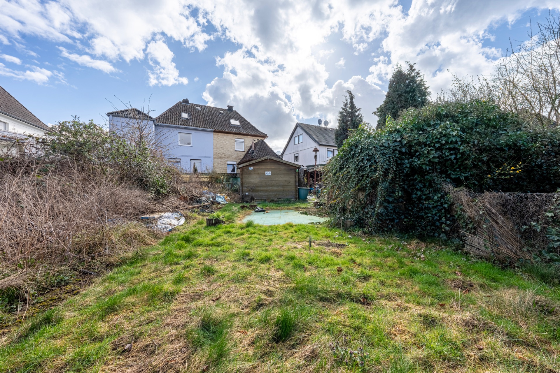 Garten mit Rasenfläche, etwas überwuchert, im Hintergrund Wohnhäuser und ein kleines Gartenhaus.