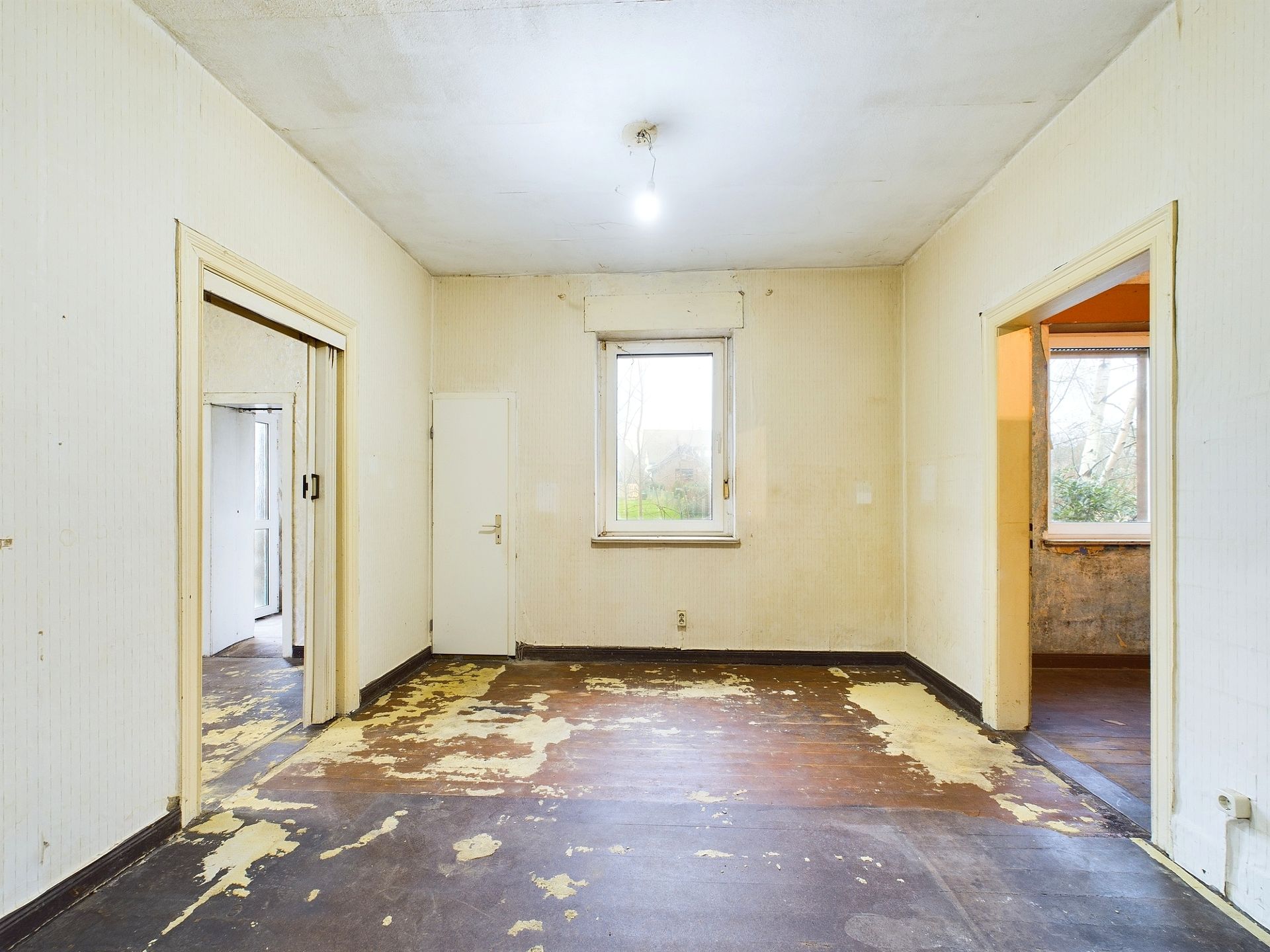 Leerer Raum mit abgenutztem Boden, einer Glühbirne an der Decke und einem Fenster an der Rückwand.
