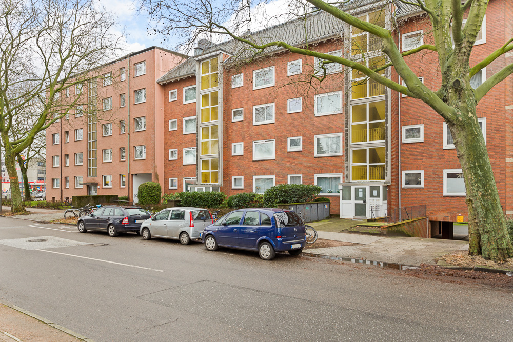 Mehrstöckiges Wohngebäude aus Backstein mit geparkten Autos und Bäumen an der Straße.