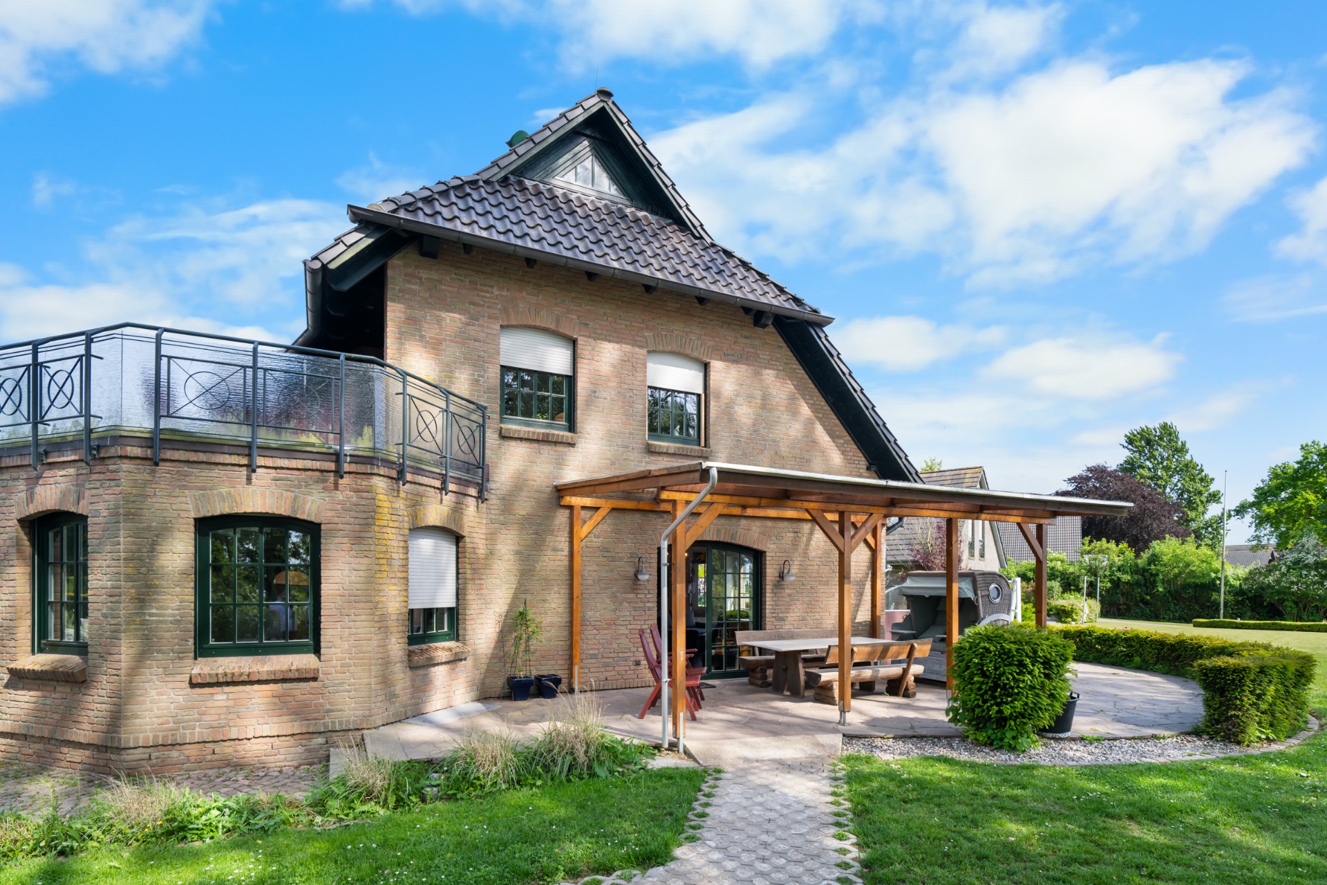 Backsteinwohnhaus mit Balkon, überdachter Terrasse und Garten bei sonnigem Wetter.