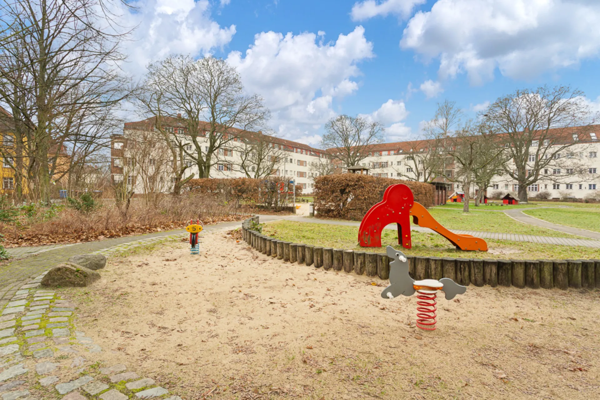 Spielplatz mit Rutsche und Wipptieren im Sand, umgeben von Wohnhäusern und Bäumen.