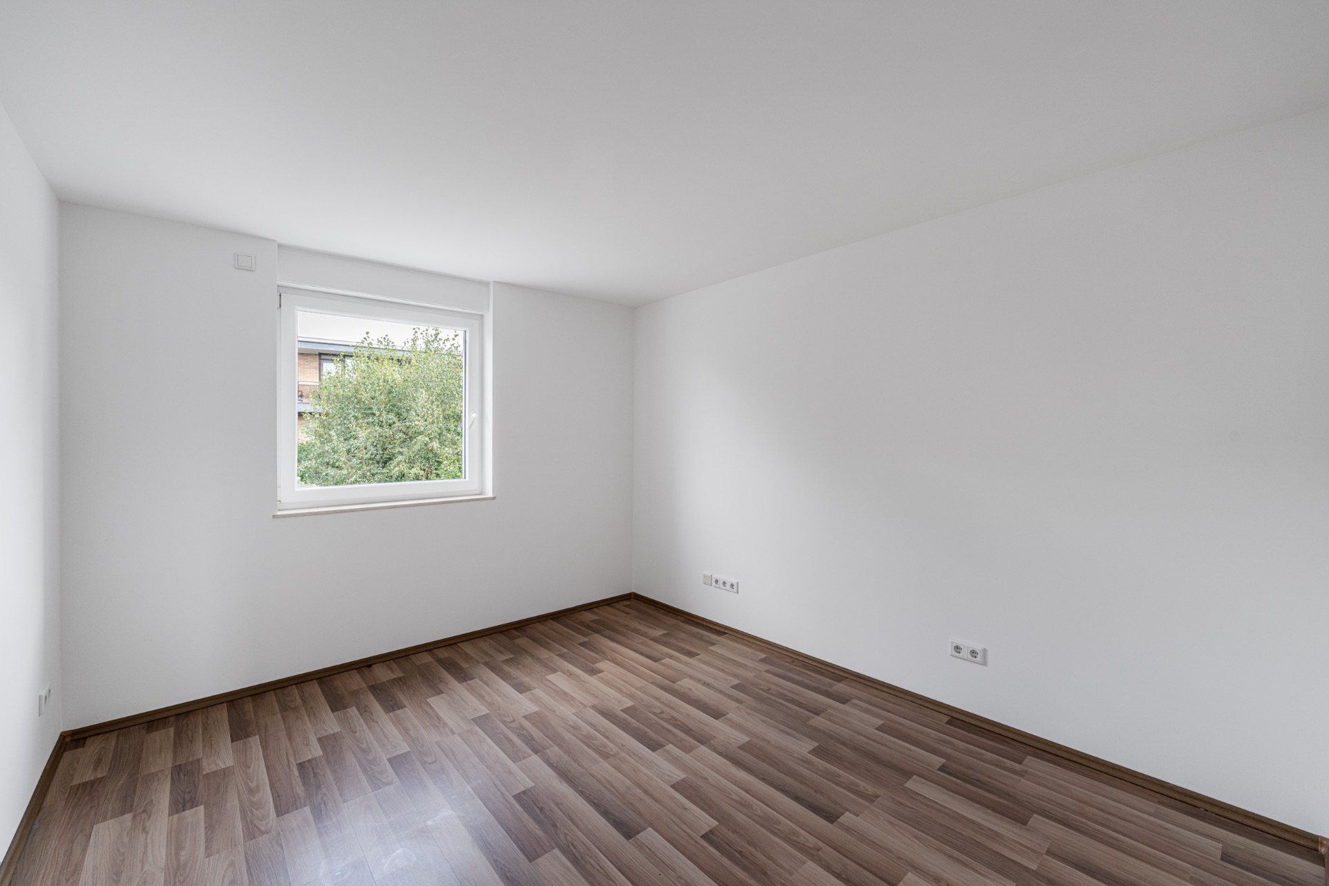 Leerer, heller Raum mit weißer Wand, großem Fenster und dunklem Holzboden.