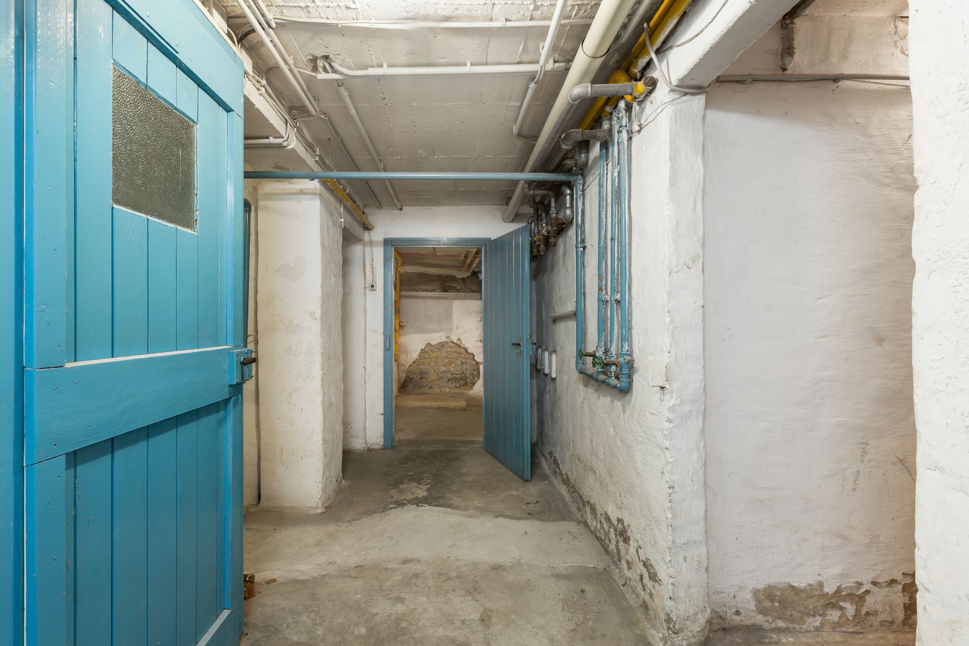 Blick in einen alten Kellerflur mit offenen blauen Türen und sichtbaren Rohrleitungen an der Decke.
