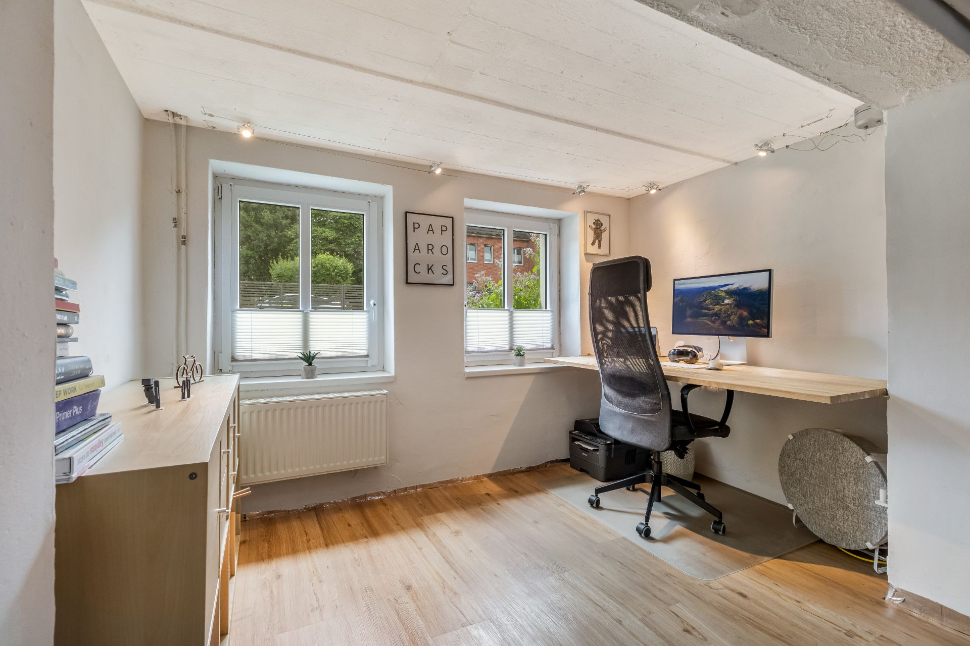 Helles Arbeitszimmer mit Schreibtisch, Bürostuhl, Regal und zwei Fenstern.