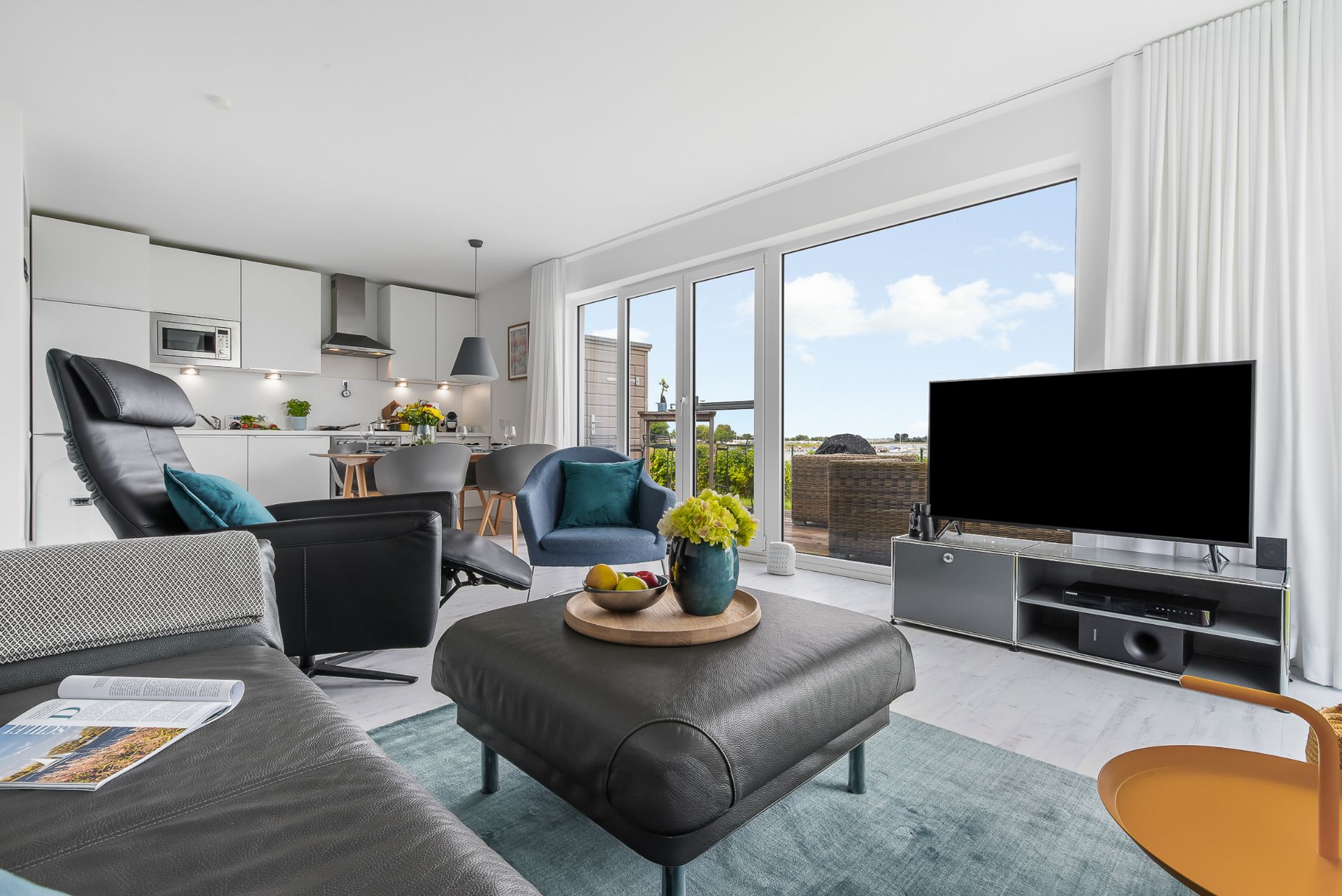 Modernes Wohnzimmer mit Leder-Sofa, TV, großen Fenstern und offener Küche.