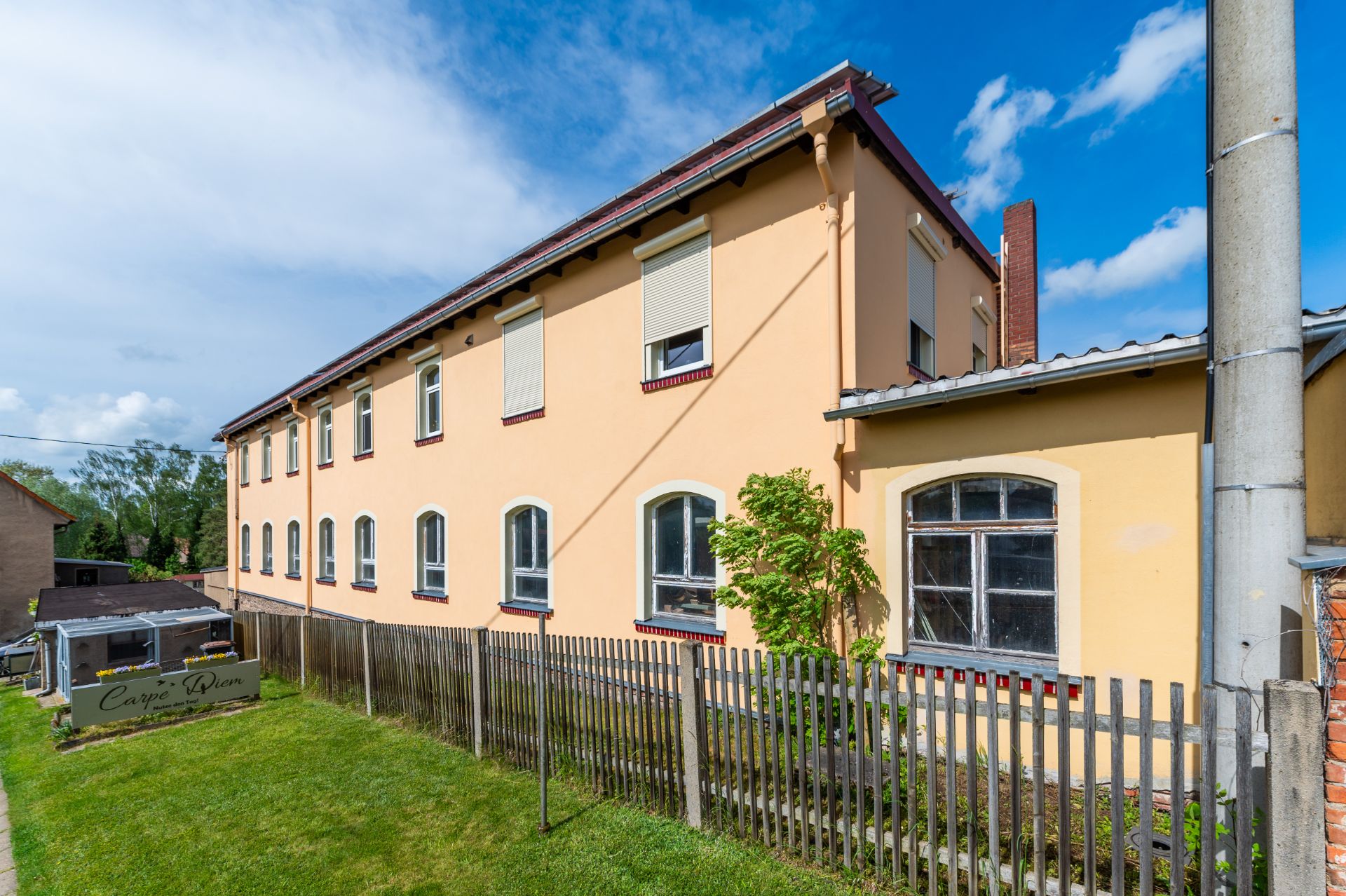 Zweistöckiges gelbes Wohnhaus mit Zaun und kleinem Garten im Vordergrund.