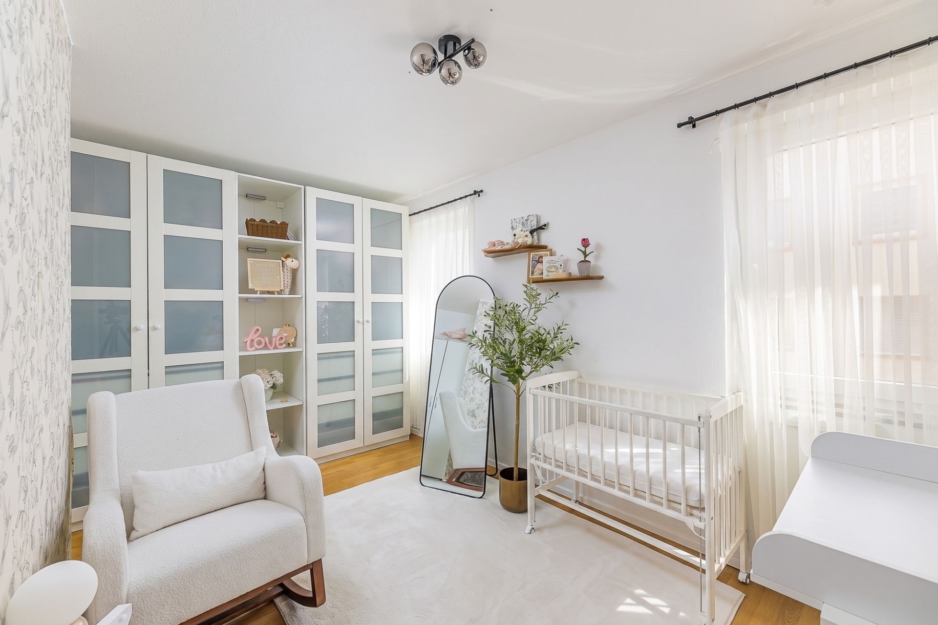 Helles Kinderzimmer mit Sessel, Gitterbett, Wickeltisch, großem Schrank und Stehspiegel.