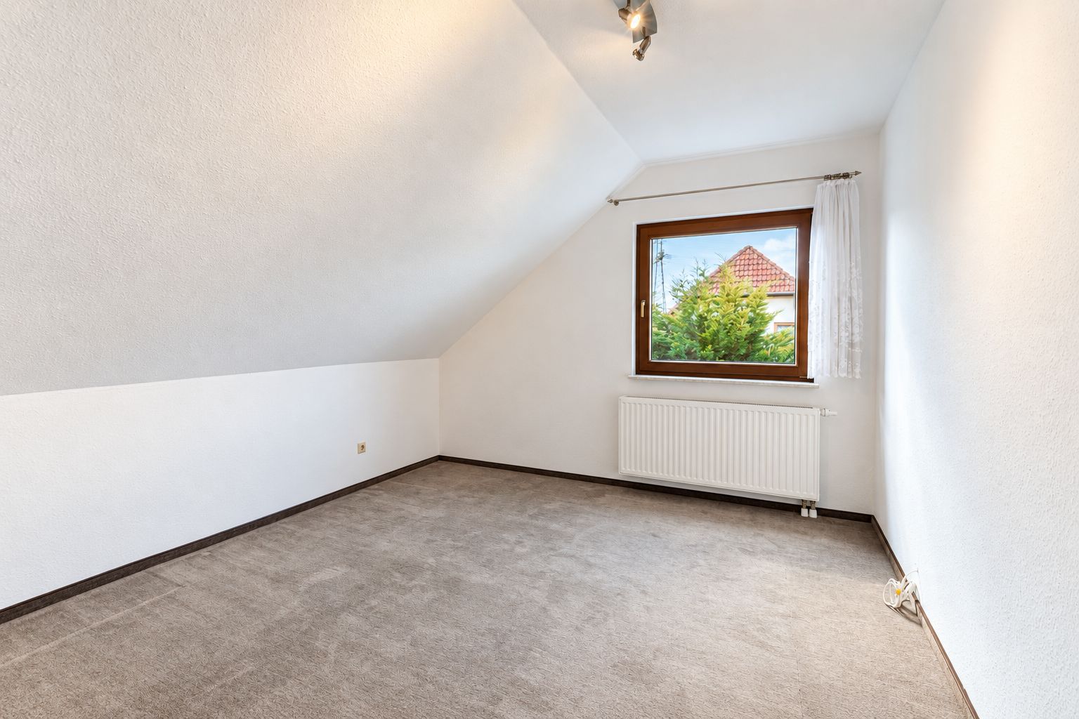 Leeres Zimmer mit Teppichboden, Dachschräge und Fenster mit Blick ins Grüne.
