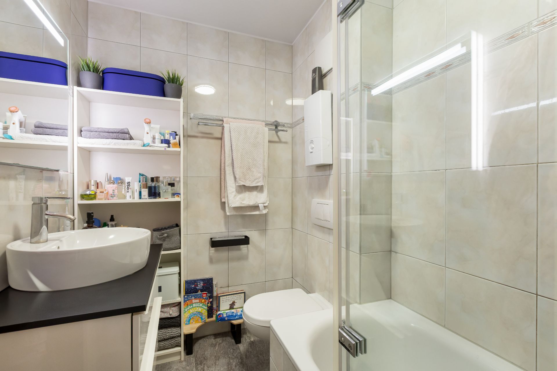 Modernes Badezimmer mit Waschbecken, Toilette, Badewanne und Regal mit Handtüchern und Pflegeprodukten.