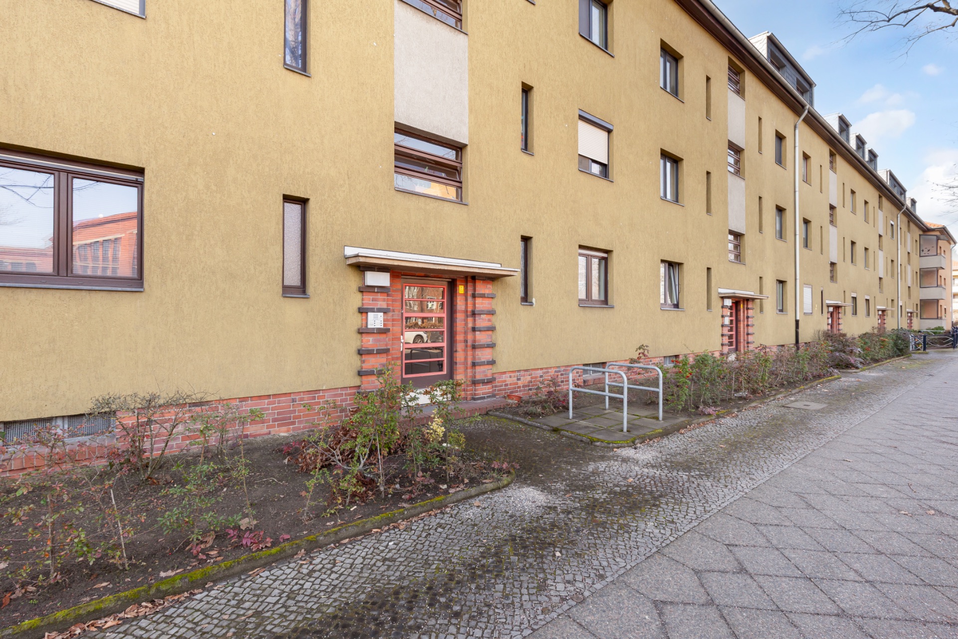 Mehrstöckiges Wohnhaus mit gelber Fassade, Fenstern und kleinen Gärten am Eingang.