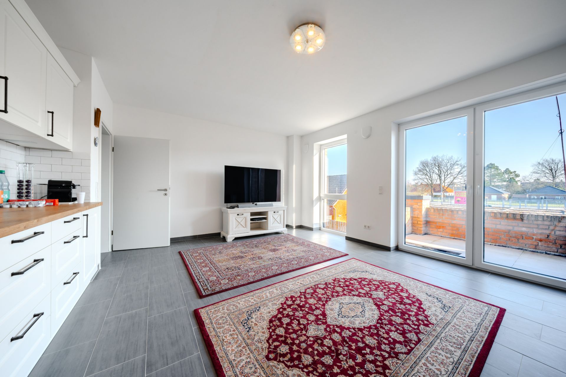 Helles Wohnzimmer mit zwei Teppichen, Fernseher, Küchenzeile und großer Fensterfront zum Balkon.